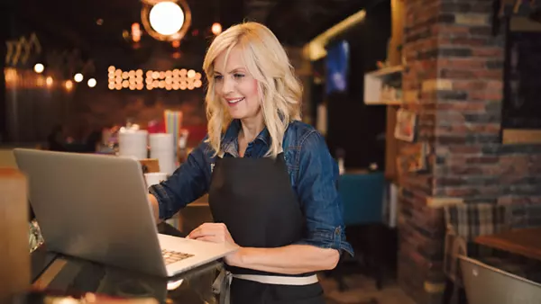 A restaurant worker reviews a laptop while standing at a countertop. 